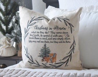 May include: A beige throw pillow with the text "Christmas in Heaven" and a poem. The design includes a Christmas tree and a wreath of leaves. The pillow is on a bed with white bedding.