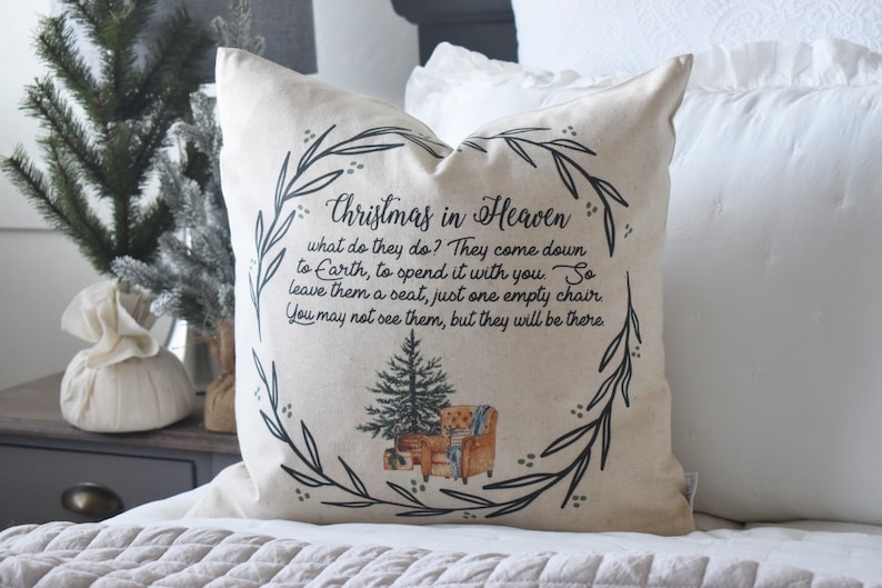 May include: A beige throw pillow with the text "Christmas in Heaven" and a poem. The design includes a Christmas tree and a wreath of leaves. The pillow is on a bed with white bedding.