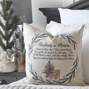 May include: A beige throw pillow with the text "Christmas in Heaven" and a poem. The pillow features a wreath design with a Christmas tree and a chair illustration. The pillow is on a bed with white bedding.