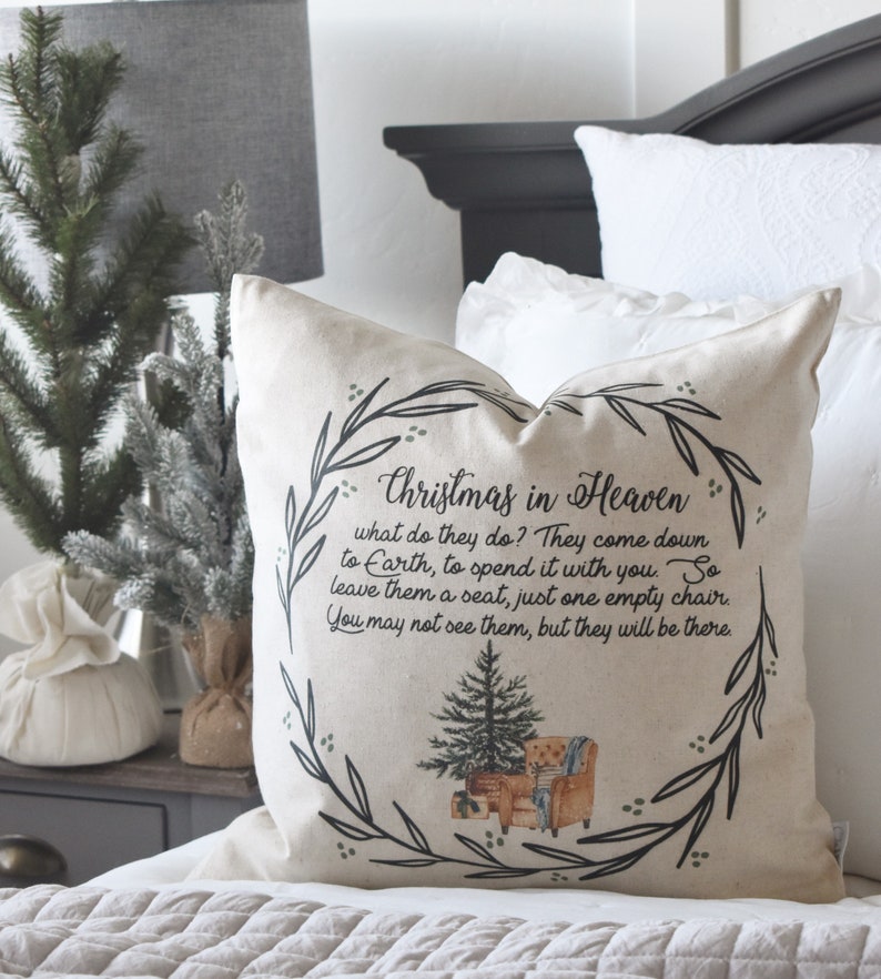 May include: A beige throw pillow with the text "Christmas in Heaven" and a poem. The pillow features a wreath design with a Christmas tree and a chair illustration. The pillow is on a bed with white bedding.