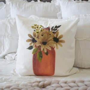 May include: A square white pillow with a watercolor design of flowers in a jar. The flowers are shades of brown and yellow, with green leaves. The jar is orange. The pillow is on a white bed.