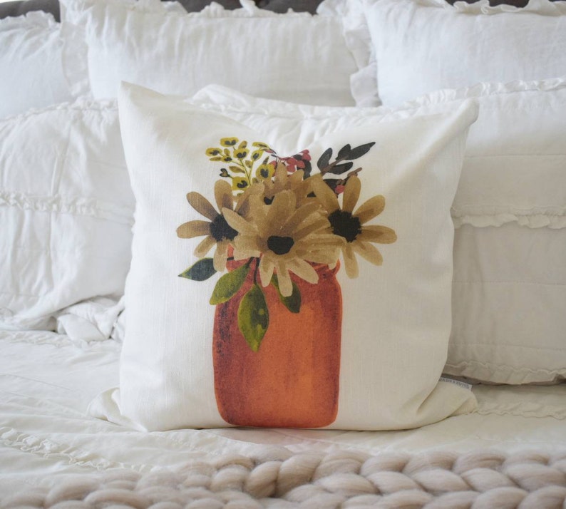 May include: A square white pillow with a watercolor design of flowers in a jar. The flowers are shades of brown and yellow, with green leaves. The jar is orange. The pillow is on a white bed.