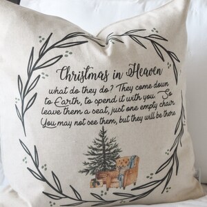 May include: A beige square pillow with the text "Christmas in Heaven" and a poem. The design includes a wreath of leaves, a Christmas tree, and a watercolor illustration of a chair. The pillow is on a white bed.