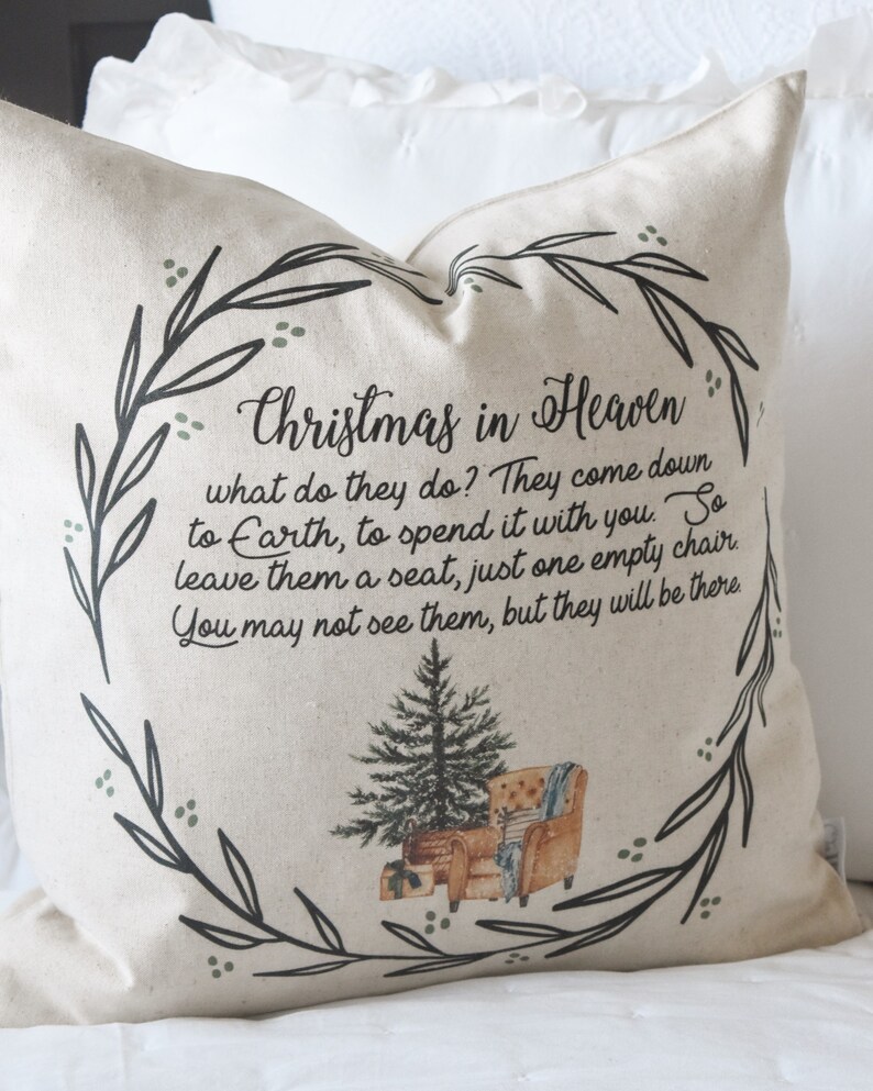 May include: A beige square pillow with the text "Christmas in Heaven" and a poem. The design includes a wreath of leaves, a Christmas tree, and a watercolor illustration of a chair. The pillow is on a white bed.