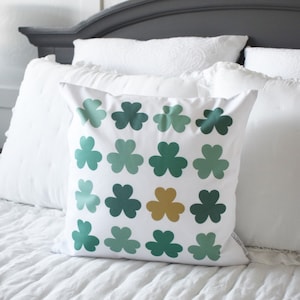 May include: A white pillow with a pattern of green shamrocks. The shamrocks are in different shades of green and there is one gold shamrock in the center.
