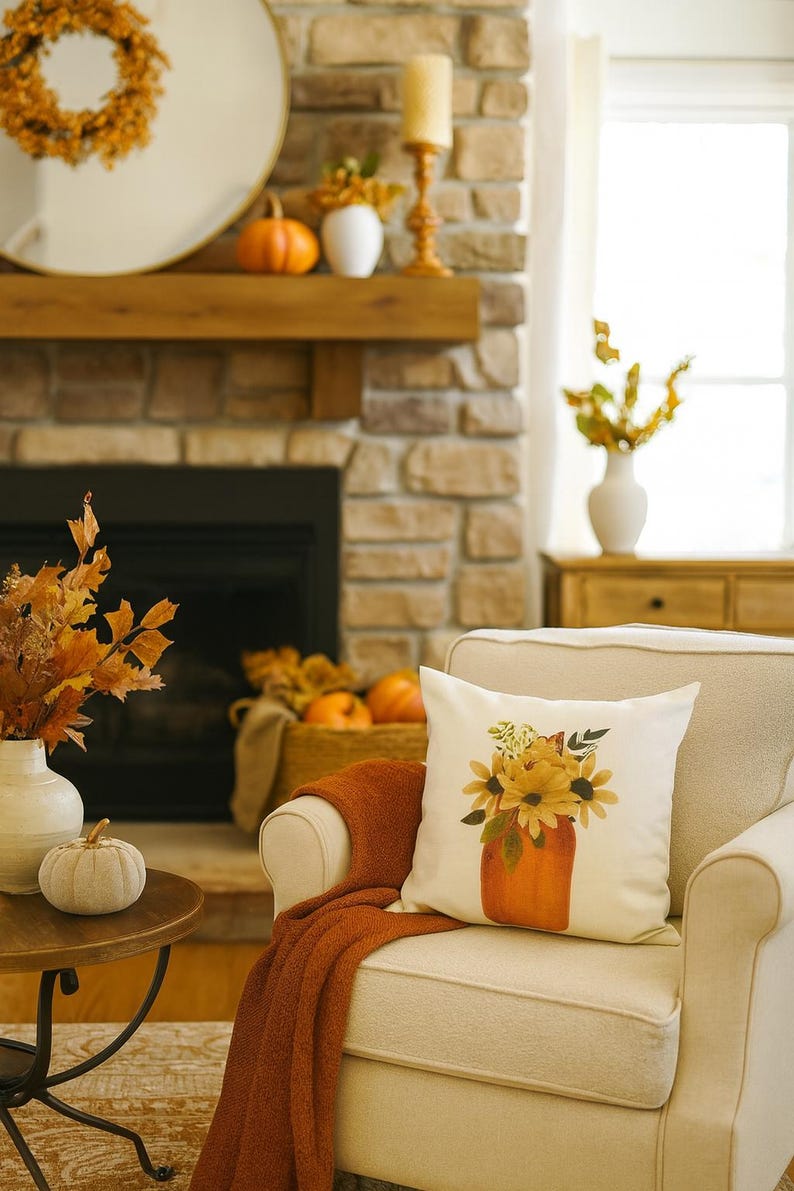May include: A cozy living room scene with fall decor. A white pillow with a sunflower design sits on a cream-colored armchair. An orange throw blanket is draped over the chair. Pumpkins, vases, and a wreath adorn the mantel.