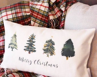 May include: A rectangular white pillow with four illustrated Christmas trees and the words "Merry Christmas" in black script. The pillow is on a gray couch with a red and green plaid blanket.