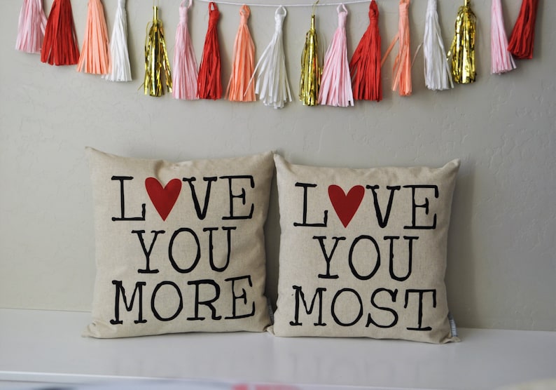 May include: Two beige throw pillows with the text "LOVE YOU MORE" and "LOVE YOU MOST" printed in black with a red heart in the middle of each pillow.