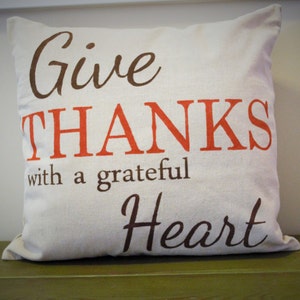 May include: Cream-colored decorative pillow with the words "Give THANKS with a grateful Heart" printed in brown and orange lettering. The pillow is square and sits on a green surface.