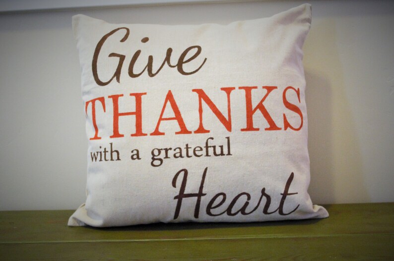 May include: Cream-colored decorative pillow with the words "Give THANKS with a grateful Heart" printed in brown and orange lettering. The pillow is square and sits on a green surface.