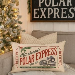 May include: A rectangular pillow with a cream background and the words "All aboard! POLAR EXPRESS TRAIN RIDE" in red and green. The pillow has a vintage train illustration and the word "BELIEVE" on each side. A Christmas tree is in the background.