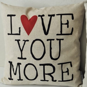 May include: A beige throw pillow with the words "I LOVE YOU MORE" printed in black with a red heart in between the words "I" and "LOVE".