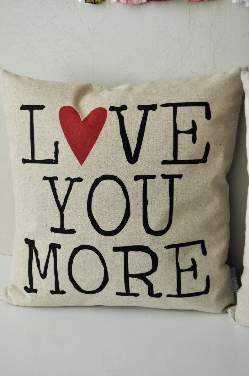 May include: A beige throw pillow with the words "I LOVE YOU MORE" printed in black with a red heart in between the words "I" and "LOVE".