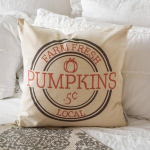 May include: A square beige pillow with a circular design featuring the words "FARM FRESH PUMPKINS" in red and brown. The design includes a pumpkin graphic and the text ".5¢ LOCAL". The pillow is on a white bed.