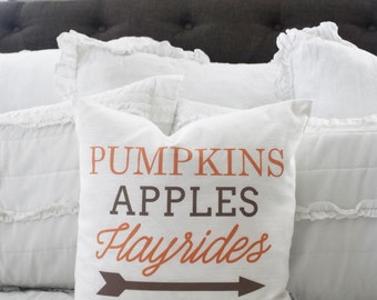 May include: A white linen pillow with the words "PUMPKINS APPLES Hayrides" in orange and brown lettering, with a brown arrow pointing right. The pillow is on a bed with white bedding.