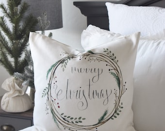 May include: A square, off-white pillow with a watercolor wreath design and the words "merry christmas" in elegant script. The wreath features green leaves, red berries, and pine needles. The pillow is on a white bed.