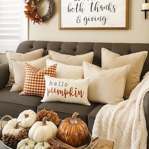 May include: A cozy living room scene with a gray sofa adorned with beige and patterned pillows. A wooden sign reads "Let our lives be full of both thanks & giving." A metal tray holds pumpkins and pinecones.