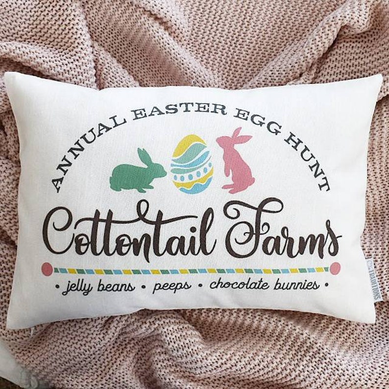 May include: White pillow with the text "ANNUAL EASTER EGG HUNT Cottontail Farms" in black script font. The pillow also features a green bunny, a pink bunny, and a colorful Easter egg. The text "jelly beans. peeps. chocolate bunnies." is printed below the main text.