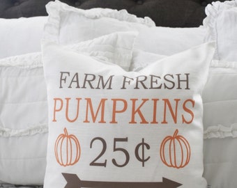 May include: A white decorative pillow with the words "FARM FRESH PUMPKINS" in brown and orange. The pillow also features two pumpkin illustrations, the price "25¢", and a brown arrow.