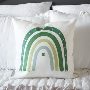 May include: A white pillow with a green, yellow, and blue rainbow design. A four-leaf clover is in the center of the rainbow.