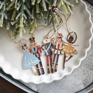 May include: Five wooden Christmas ornaments depicting characters from the Nutcracker ballet. The ornaments are painted in bright colors and feature a ballerina, a nutcracker soldier, a sugar plum fairy, and two other nutcracker soldiers.