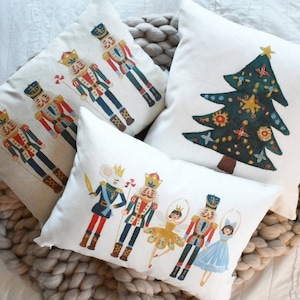 May include: Four white decorative throw pillows with holiday-themed illustrations. One pillow features a green Christmas tree with gold stars and ornaments. The other pillows have nutcracker soldiers and ballet dancers in colorful outfits.
