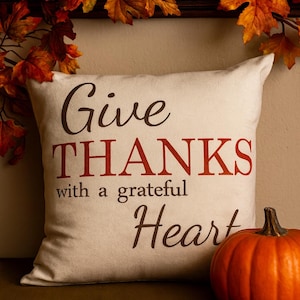 May include: Cream-colored decorative pillow with the words "Give THANKS with a grateful Heart" in brown and red lettering. A small orange pumpkin sits to the right. Autumn leaves and pumpkins decorate the background.