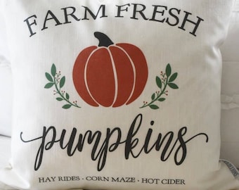 May include: Cream-colored decorative pillow with the words "FARM FRESH PUMPKINS" in black script. A large orange pumpkin graphic is centered on the pillow, with green leaf accents. The bottom of the pillow reads "HAY RIDES • CORN MAZE • HOT CIDER."