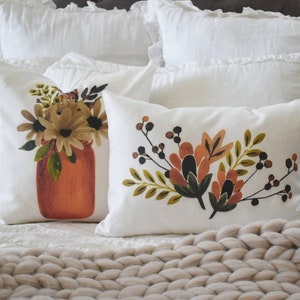 May include: Two white throw pillows with floral designs. One pillow features a bouquet in a rust-colored jar. The other pillow has a floral arrangement with orange, brown, and green leaves and berries. The pillows are on a white bed.