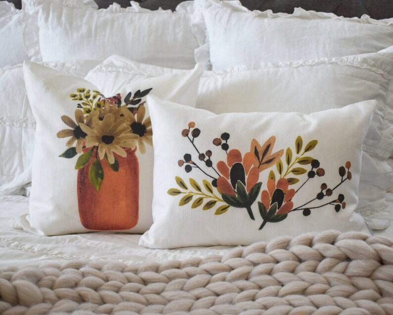 May include: Two white throw pillows with floral designs. One pillow features a bouquet in a rust-colored jar. The other pillow has a floral arrangement with orange, brown, and green leaves and berries. The pillows are on a white bed.