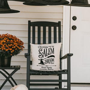 May include: A black rocking chair with a pillow that reads "Salem Broom Co." is on a porch decorated for Halloween. Black witch hats hang above. Pumpkins and orange mums complete the fall scene.