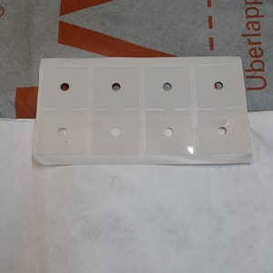 Puede incluir: Una hoja de papel blanca con seis cuadrados de plástico transparente, cada uno con un pequeño agujero en el centro. Los cuadrados están dispuestos en dos filas de tres. El texto "Überlappung" es visible en la esquina superior derecha de la imagen.