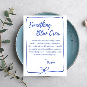 以下が含まれることがあります： 水色のプレートに「Something Blue Crew」と書かれた青い文字の白いカード。カードにはメッセージと「Brianna」という名前が書かれています。青いリボンのグラフィックが下にあります。