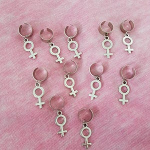 Puede incluir: Diez pinzas para el pelo plateadas con un dije en forma de s&iacute;mbolo femenino que cuelga de cada pinza. Las pinzas est&aacute;n dispuestas en forma escalonada sobre un fondo rosa.