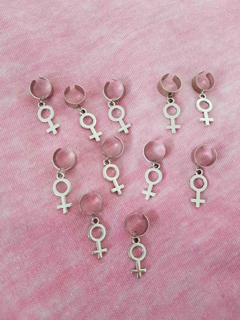 Puede incluir: Diez pinzas para el pelo plateadas con un dije en forma de s&iacute;mbolo femenino que cuelga de cada pinza. Las pinzas est&aacute;n dispuestas en forma escalonada sobre un fondo rosa.