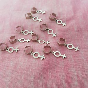 Puede incluir: Un conjunto de diez pinzas para el pelo plateadas con un dije de s&iacute;mbolo femenino que cuelga de cada pinza. Las pinzas est&aacute;n dispuestas en semic&iacute;rculo sobre un fondo de tela rosa.