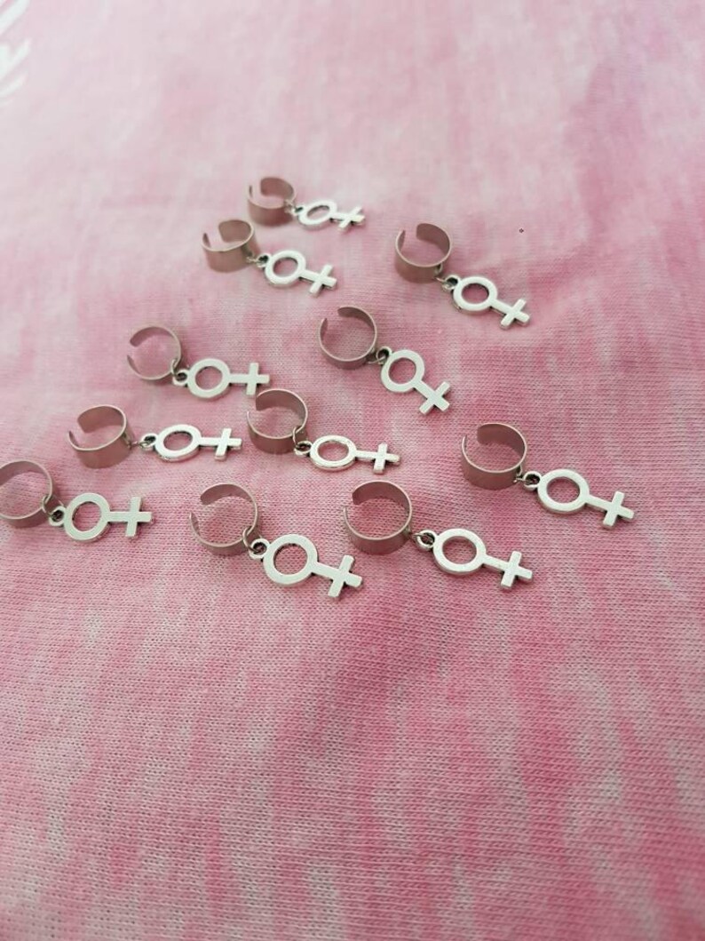 Puede incluir: Un conjunto de diez pinzas para el pelo plateadas con un dije de s&iacute;mbolo femenino que cuelga de cada pinza. Las pinzas est&aacute;n dispuestas en semic&iacute;rculo sobre un fondo de tela rosa.