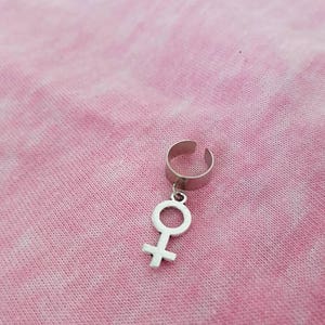 Puede incluir: Un pendiente de aro plateado con un colgante en forma de s&iacute;mbolo femenino.