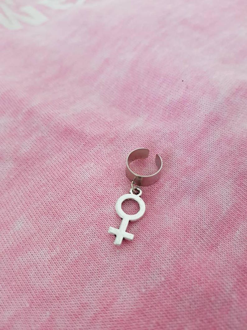 Puede incluir: Un pendiente de aro plateado con un colgante en forma de s&iacute;mbolo femenino.