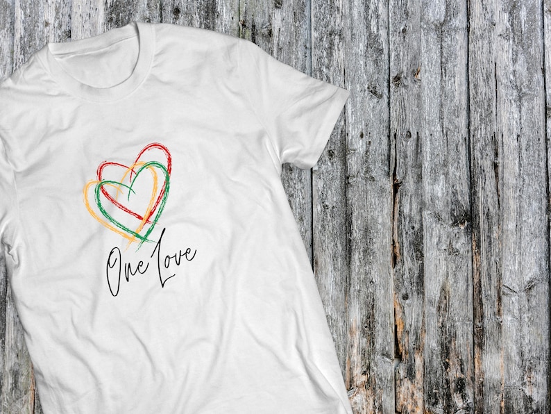 May include: White t-shirt with a green, yellow, and red heart design and the text "One Love" in black script.