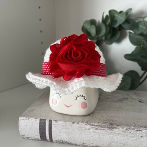 May include: A ceramic marshmallow figurine with a smiling face, wearing a white crocheted hat adorned with a red fabric flower and a red polka dot ribbon. The figurine is resting on a book.