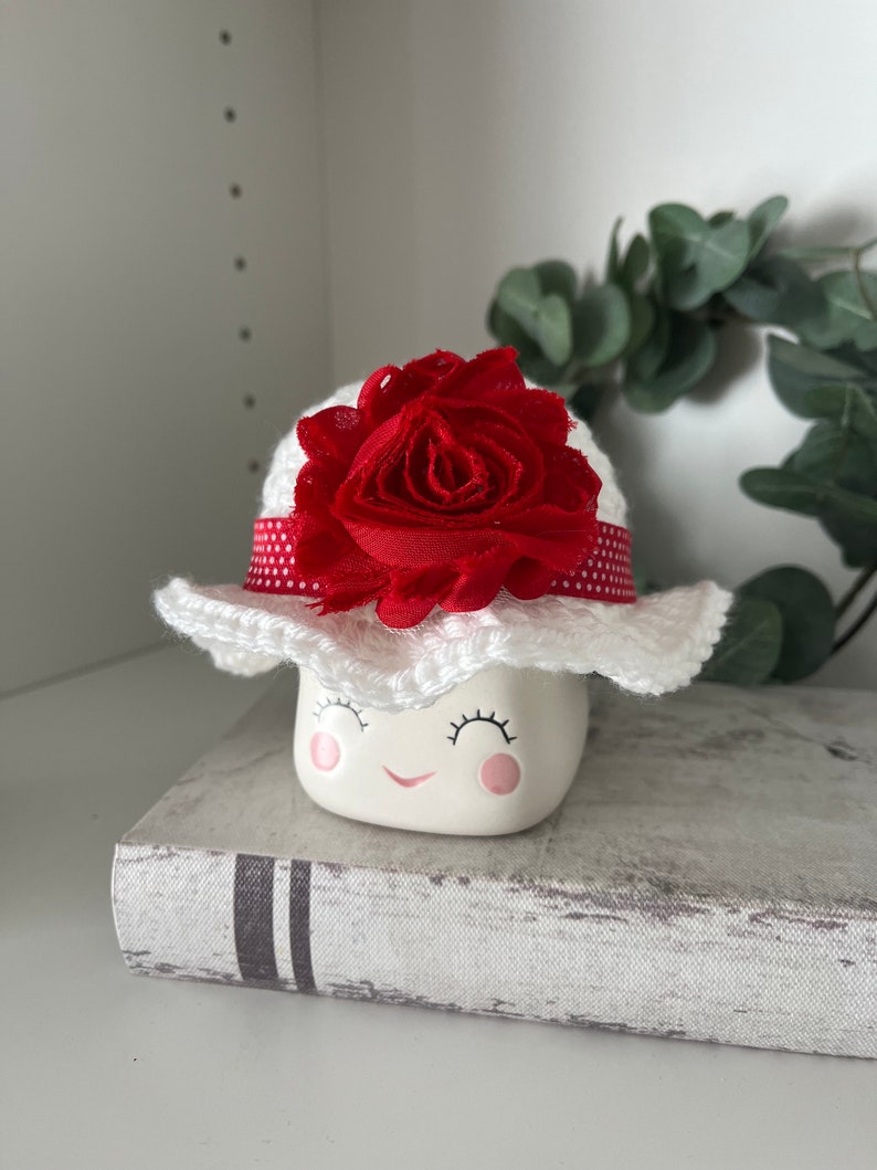 May include: A ceramic marshmallow figurine with a smiling face, wearing a white crocheted hat adorned with a red fabric flower and a red polka dot ribbon. The figurine is resting on a book.