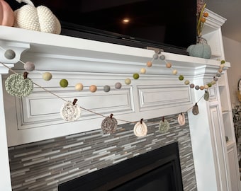 Puede incluir: Guirnalda decorativa con calabazas de ganchillo y bolas de fieltro en tonos otoñales de verde, marrón y crema. La guirnalda está ensartada en hilo de yute y se exhibe sobre una chimenea, creando un ambiente acogedor.