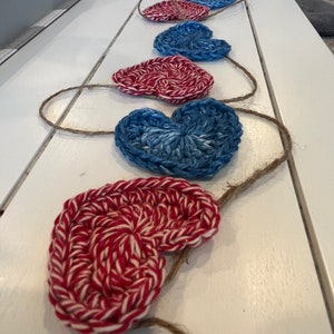 May include: A garland of crocheted hearts in red and white, and blue and white, strung on a natural twine. The hearts are arranged in a row, creating a decorative accent. The garland is placed on a white surface.