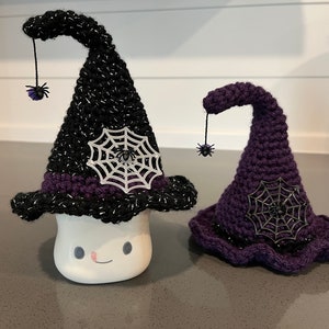 May include: Two Halloween witch hats. One is black with silver accents and a purple band, the other is purple. Both have a spider web and a dangling spider decoration. The black hat is on a white ceramic marshmallow.