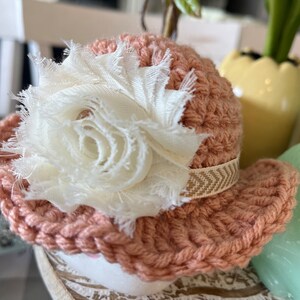 May include: A small, peach-colored crocheted hat with a white fabric flower and a beige ribbon. The hat is likely handmade and has a decorative, rustic appearance. The hat is sitting on a white surface.
