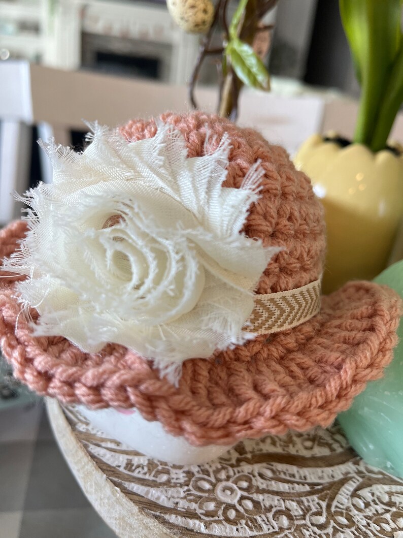May include: A small, peach-colored crocheted hat with a white fabric flower and a beige ribbon. The hat is likely handmade and has a decorative, rustic appearance. The hat is sitting on a white surface.