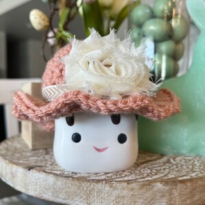 May include: A white ceramic figure with a smiling face, wearing a pink crocheted hat adorned with a cream-colored fabric flower. The figure is on a wooden tray, with a green bunny and decorative items in the background.