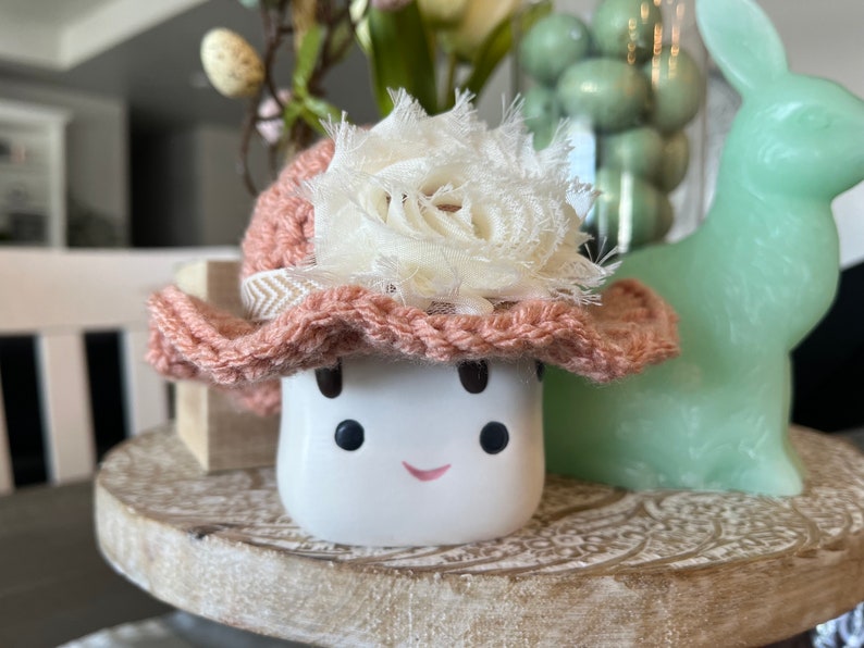 May include: A white ceramic figure with a smiling face, wearing a pink crocheted hat adorned with a cream-colored fabric flower. The figure is on a wooden tray, with a green bunny and decorative items in the background.