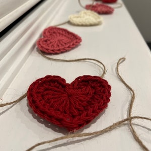 May include: A handmade garland of crocheted hearts in shades of red and cream. The hearts are strung on natural twine, creating a rustic and charming decorative piece. Perfect for Valentine's Day or adding a touch of love to any space.
